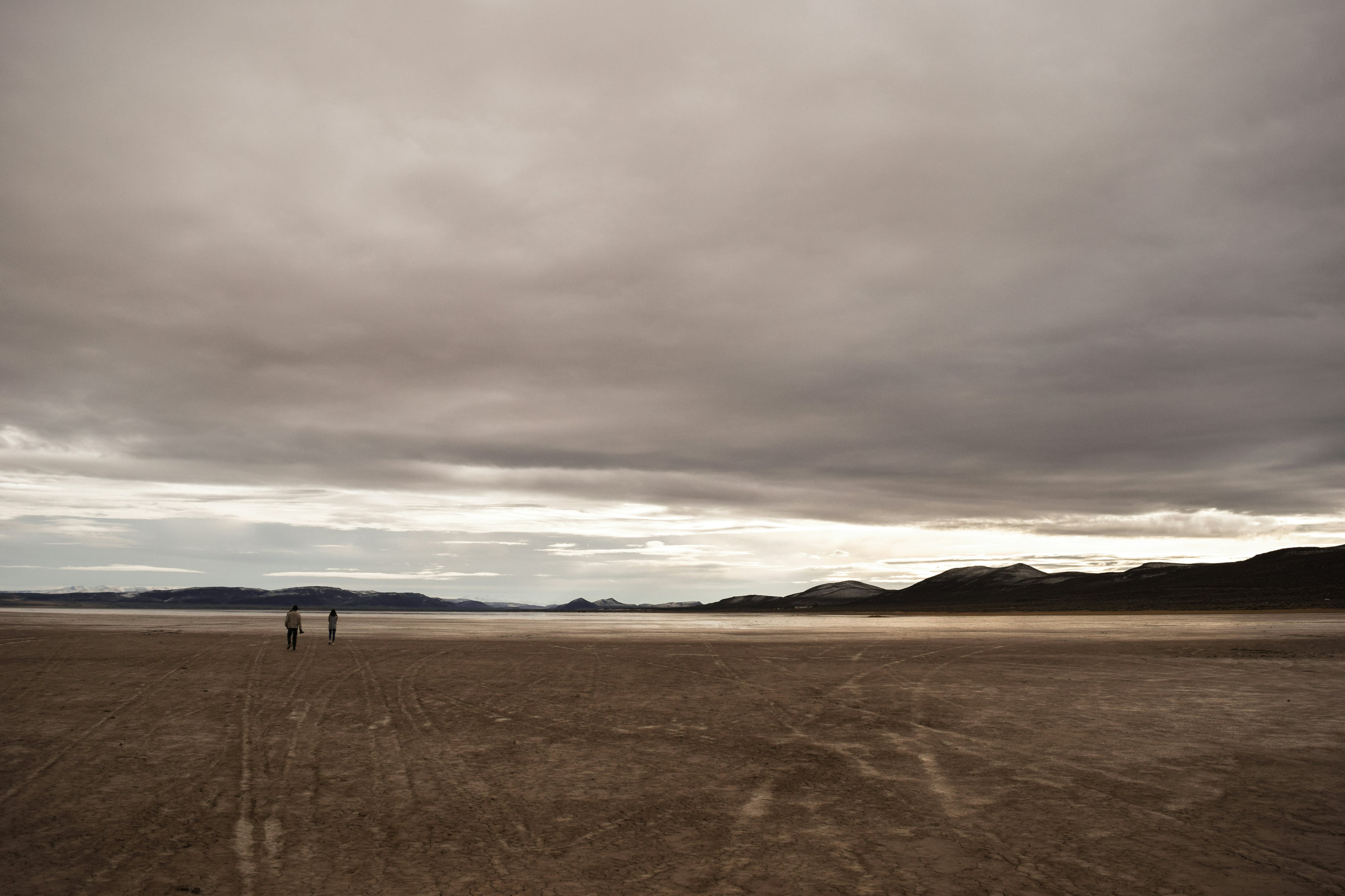 Two people walking across the Alvord Desert playa beneath a sweeping cloudy sky